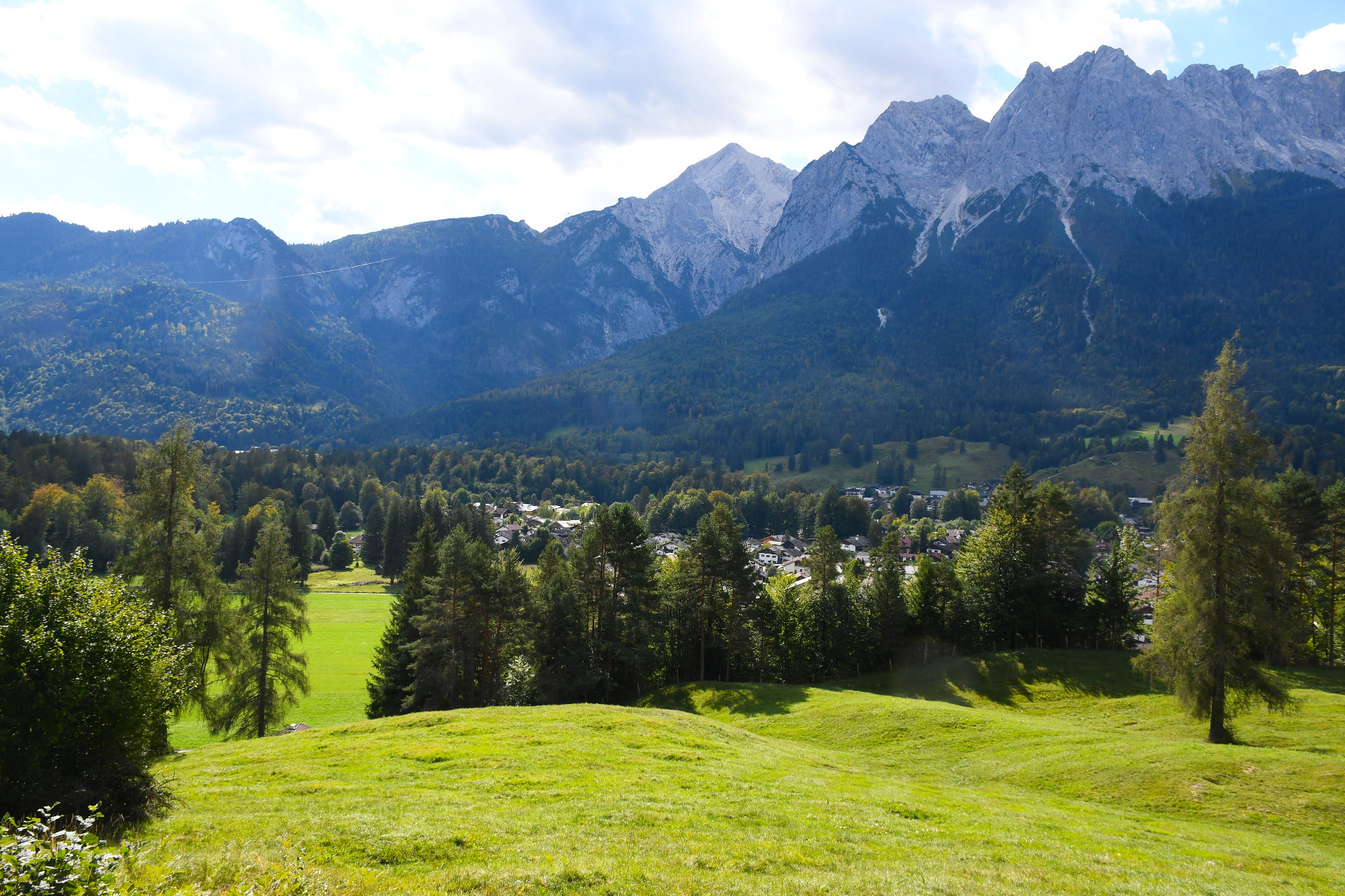 Grüne Wiese mit Holzschuppen und Bäumen vor hohen Alpenbergen unter hellem, wolkigem Himmel.